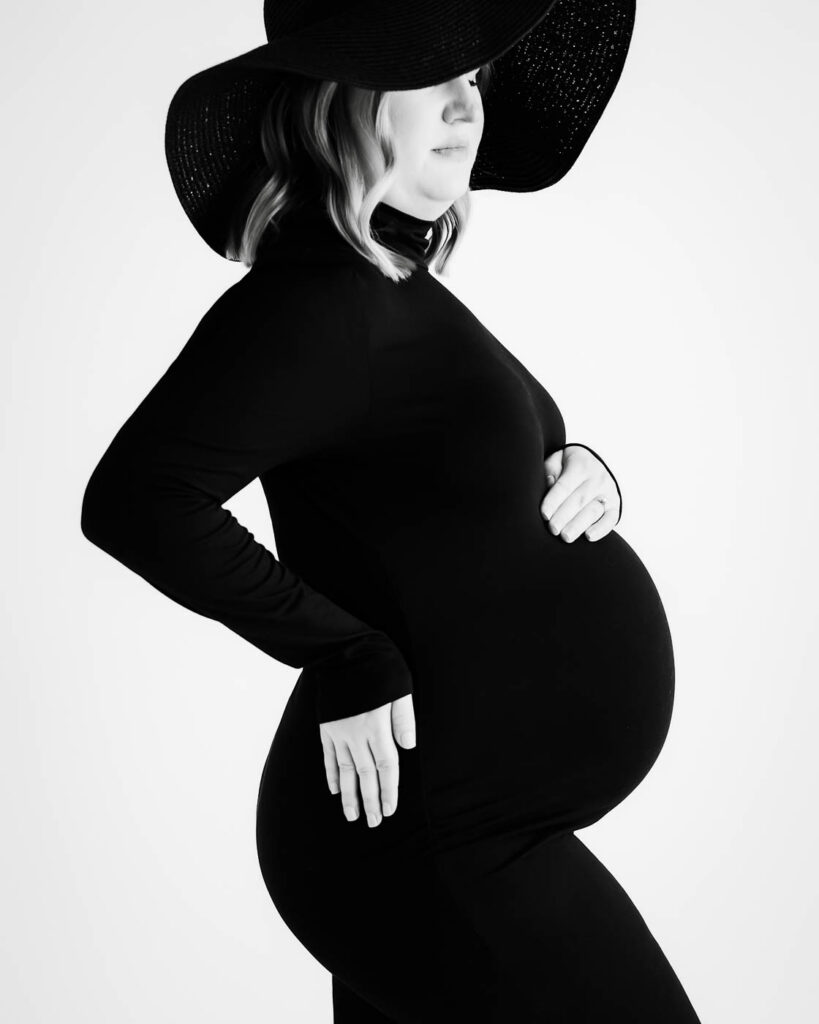 Elegant Black And White Maternity Portrait Of Expecting Mother Wearing Fitted Black Dress And Wide Brim Hat In Dallas Photography Studio.