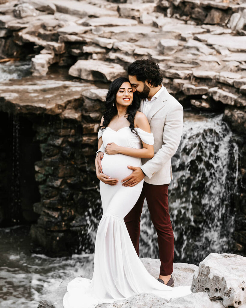 Couple Posing Beside A Waterfall During A Winter Maternity Shoot In Dallas Wearing Elegant Neutral Maternity Outfits.