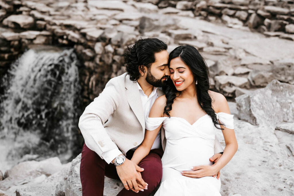 Couple Embracing Beside A Waterfall During A Winter Maternity Shoot In Dallas With Elegant Neutral Maternity Styling.