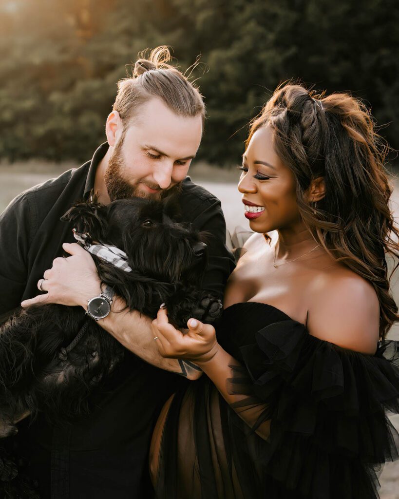 Maternity Photos With Pets: When Your First Baby Joins The Session 7 Maternity Photos With Pets In Dallas Featuring An Expecting Couple Holding Their Black Dog During An Outdoor Maternity Session At Sunset.