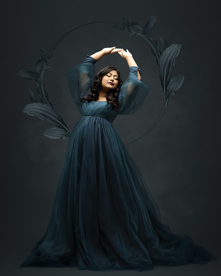 A pregnant woman in a blue dress posing in front of a black background.