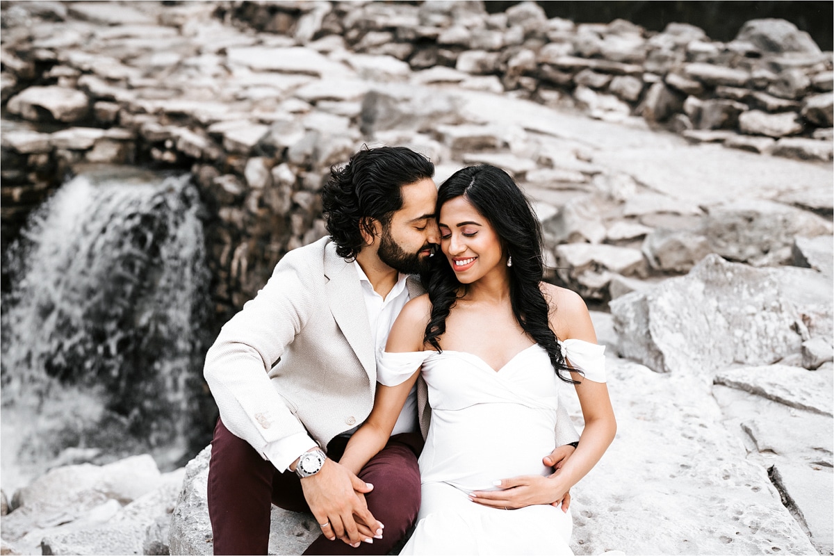 Pregnant Couple Sitting Near Waterfall Both Smiling For Dallas Pregnancy Photographer