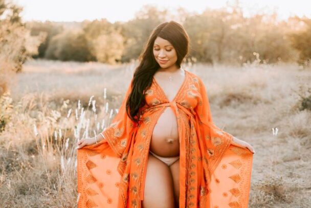  A Pregnant Woman Stands In A Field Of Wild Grass With An Orange Gown Tied Below The Breast And Open From Below, Showing Her Protruding Baby Bump And Legs During Some Of The Best Maternity Photography In Dallas.