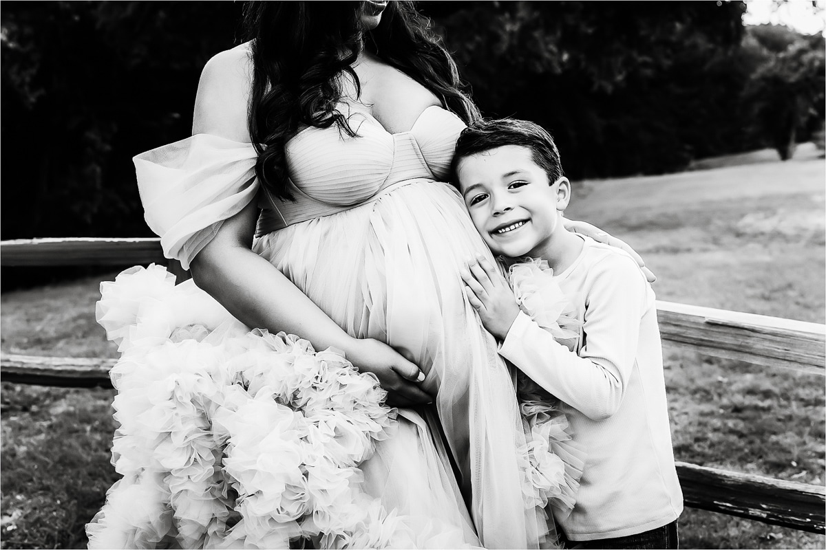 A Pregnant Woman In A Flowing Dress Cradles Her Belly While A Young Boy Stands Beside Her, Smiling And Resting His Head Against Her Stomach. This Heartwarming Moment Of Maternity Photos With Siblings Is Captured Outdoors Near A Wooden Fence.