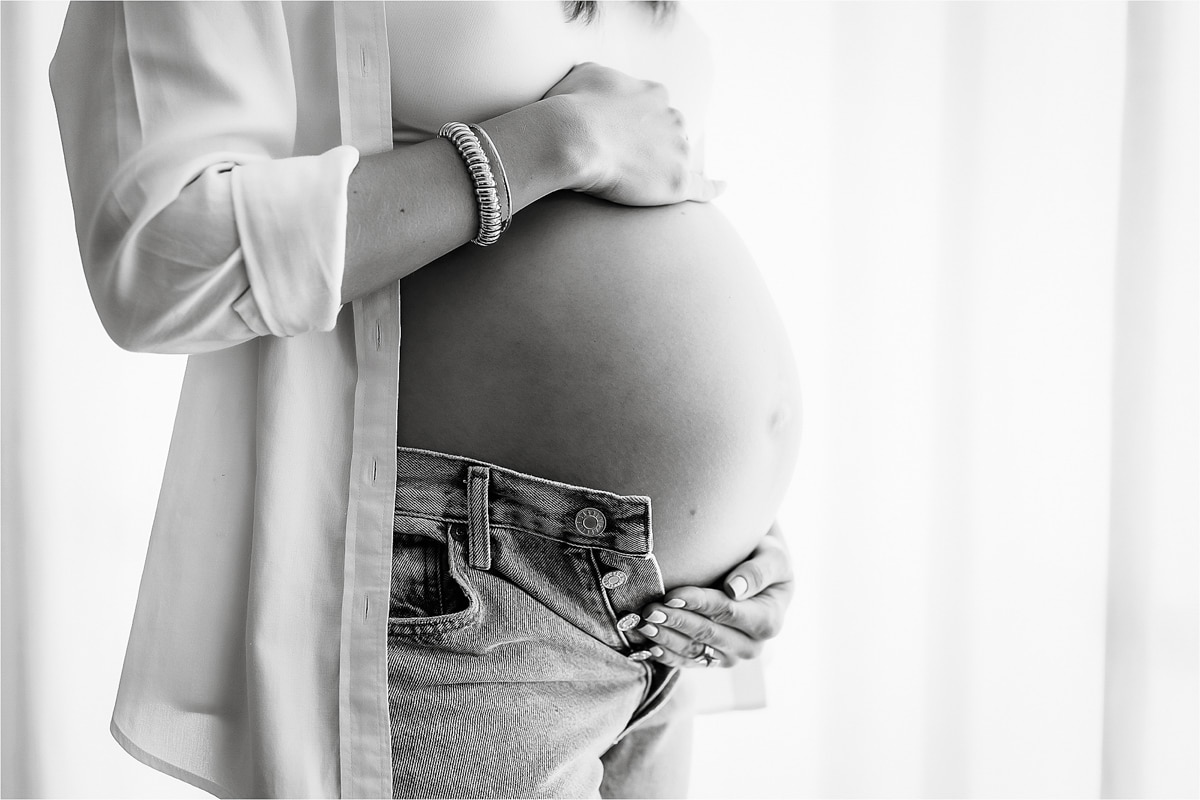 A Pregnant Person Wearing An Open Shirt And Jeans Holds Their Bare Belly With One Hand And Adjusts The Waistband With The Other, Capturing A Beautiful Moment Perfect For A Maternity Photography Business.