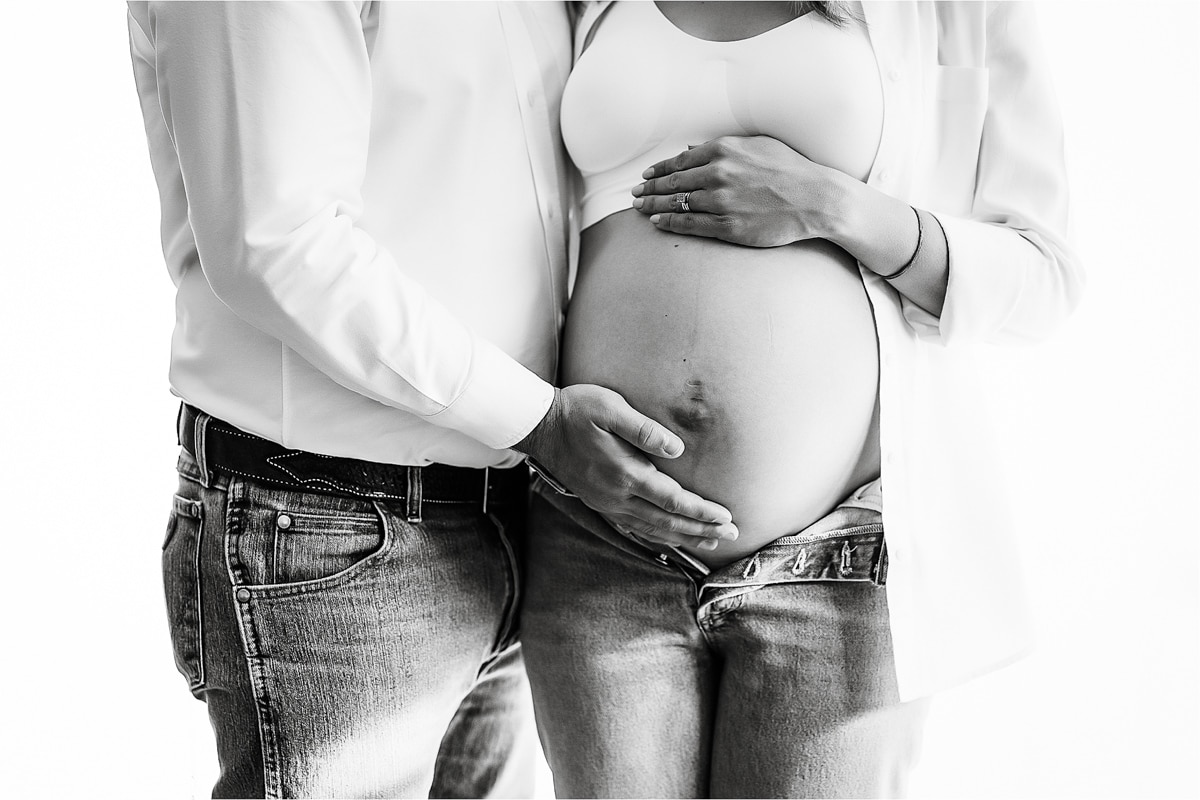 A Close-Up Of A Pregnant Couple With Their Hands Placed On The Woman'S Exposed Belly, Captured Perfectly By Our Maternity Photography Business. Both Are Wearing Casual Jeans And White Tops.