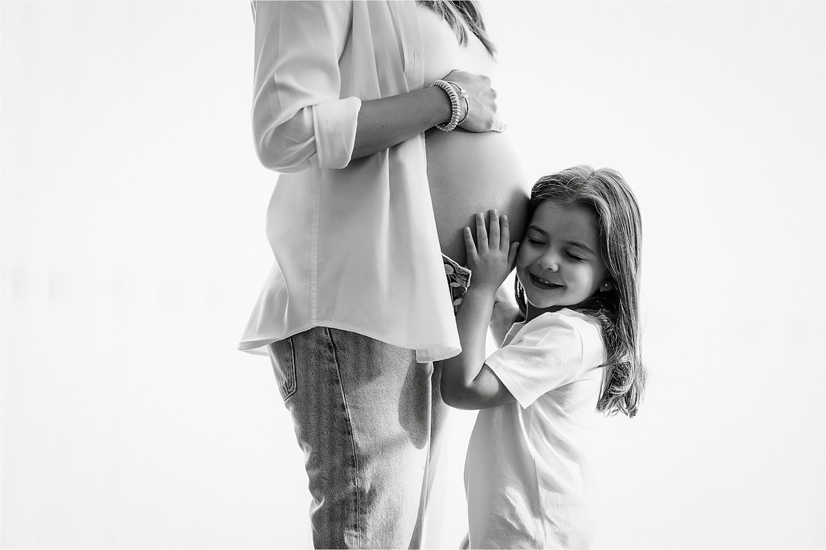 A Young Girl With Long Hair Smiles And Rests Her Head Against The Exposed Pregnant Belly Of A Woman Standing Beside Her, Perfectly Capturing A Serene Moment. Both Are Wearing Light-Colored Clothes, Embodying The Elegance Typical Of A Maternity Photography Business. The Background Is Plain And Light.