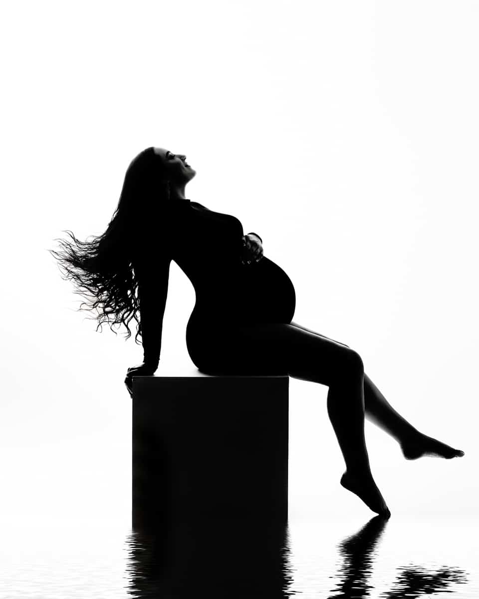 Silhouette of a pregnant woman with long hair, in a seated pose on a cube with one leg extended forward, against a bright white background.