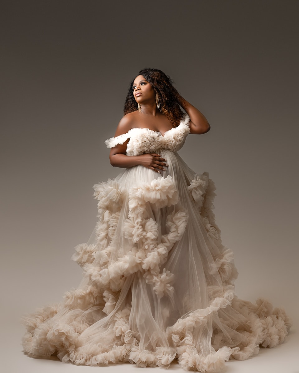 A woman stands against a neutral background, wearing a voluminous off-shoulder gown adorned with ruffles. She has one hand on her hip and the other touching her hair.