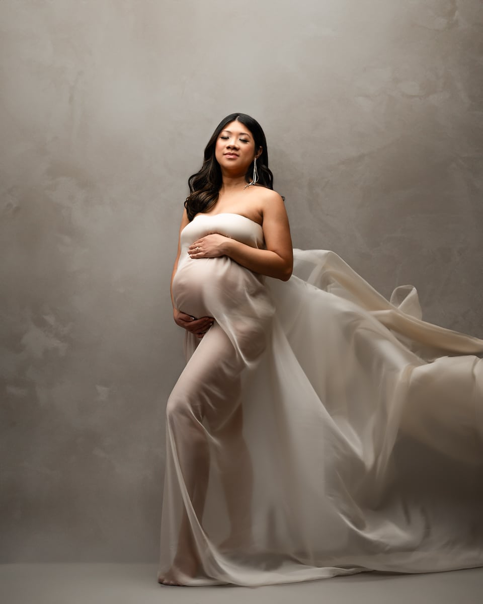 A pregnant woman stands with a flowing sheer cloth draped around her, cradling her belly while looking at the camera in a softly lit studio setting.