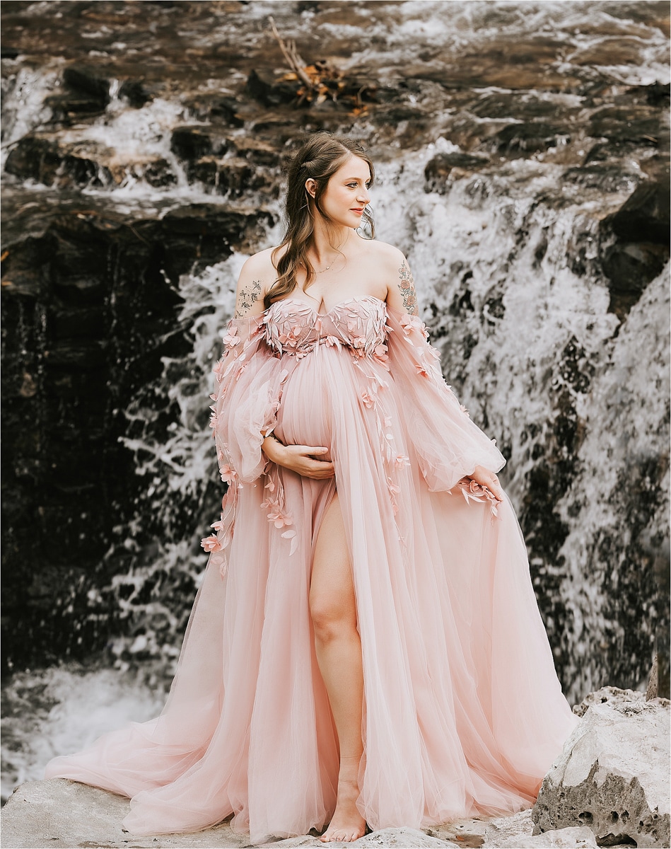 Pregnant Woman In A Flowing Pink Gown Stands In Front Of A Rocky Waterfall, Gently Holding Her Belly And Looking To The Side, Captured Beautifully By A Skilled Maternity Photographer.