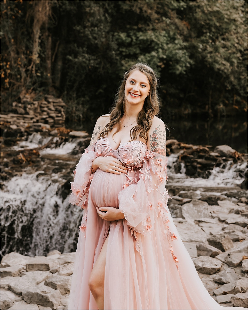 A Smiling Pregnant Woman In A Flowing Pink Dress Poses In Front Of A Waterfall And Rocky Terrain, Captured Beautifully By A Talented Maternity Photographer.