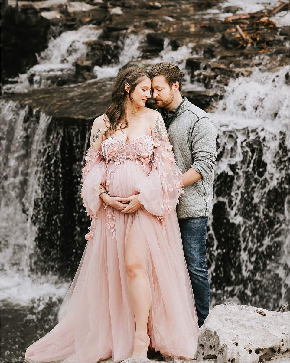 A Maternity Photographer Captures A Tender Moment As A Pregnant Woman In A Pink Dress Stands With A Man In A Gray Sweater By A Waterfall. The Man Embraces Her From Behind, Both Gazing Down Lovingly.