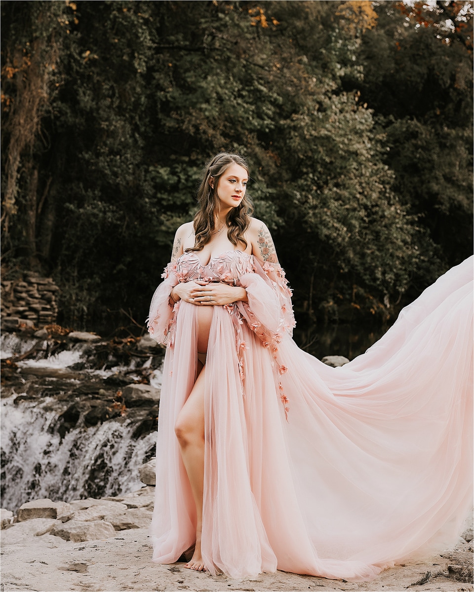 A Woman In A Flowing Pink Gown Stands In Front Of A Natural Waterfall, Surrounded By Lush Greenery, Captured Beautifully By A Skilled Maternity Photographer.