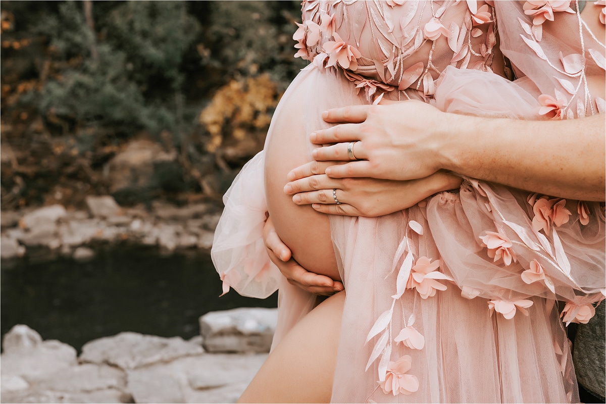 Close-Up Of A Couple Holding A Pregnant Woman'S Belly, Captured By A Skilled Maternity Photographer. The Woman Wears A Pink Dress With Floral Details, And Both Have Wedding Bands. Rocks And Greenery Are Visible In The Background.