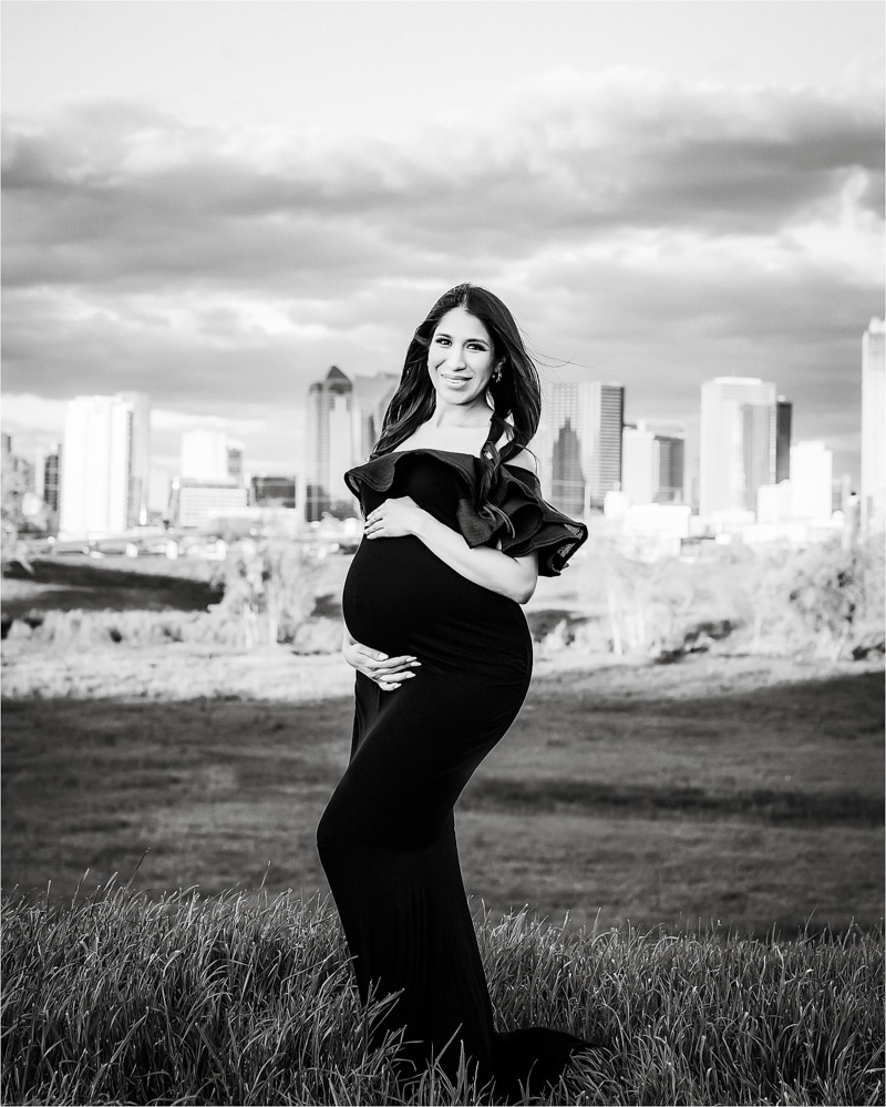 A Pregnant Woman In A Long, Black Dress Poses Outdoors With A Cityscape In The Background.