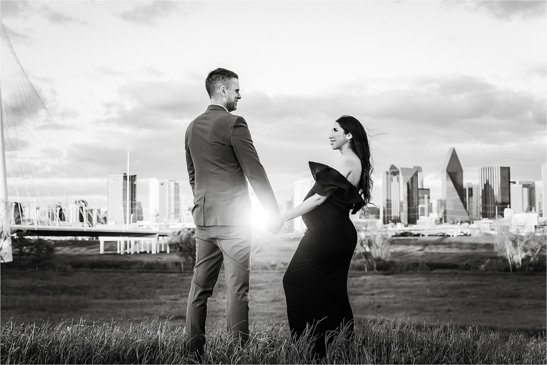 A Couple Stands Hand-In-Hand In A Grassy Field, Facing Each Other, With A City Skyline In The Background During Sunset.