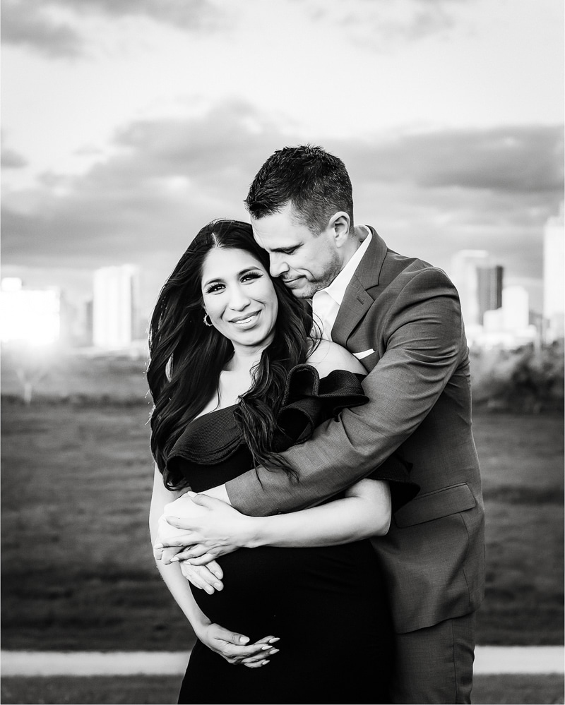 A Couple Stands Together Outdoors; The Man Embraces The Woman From Behind As She Holds Her Pregnant Belly, Both Smiling Softly. A City Skyline Is Visible In The Background Under A Cloudy Sky.