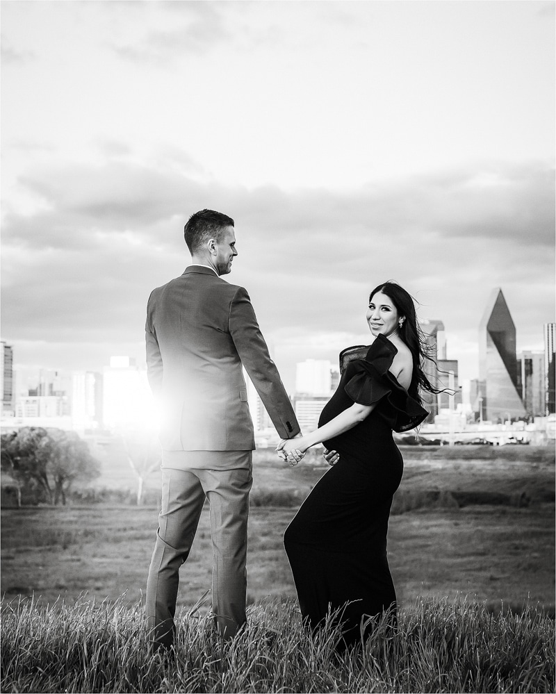 A Man In A Suit And A Woman In A Long, Black Off-The-Shoulder Dress Pose Together In A Grassy Area With A Cityscape In The Background.