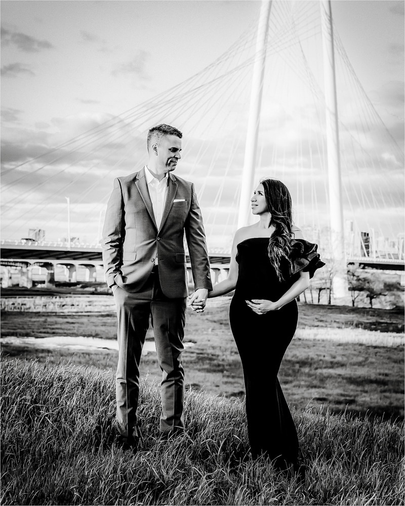 A Man And A Woman, Dressed Formally, Hold Hands And Stand On A Grassy Field In Front Of A Modern Cable-Stayed Bridge. The Woman Is Wearing An Off-The-Shoulder Dress.