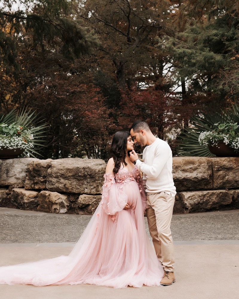 Couple Stands Outside In Front Of A Stone Wall Smiling At Each Other For Maternity Photos Dallas Arboretum