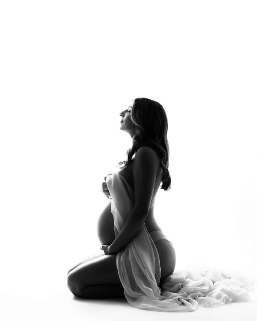 Elegant Maternity Photography That Never Feels Dated 12 Pregnant Woman Kneeling And Wrapped In Sheer Fabric During A Black And White Maternity Silhouette Session In Dallas