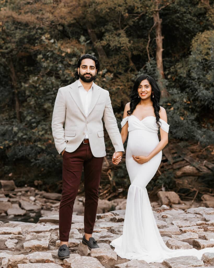 Expecting Couple Standing On Rocks Looking At Camera Smiling For A Maternity Photoshoot Dallas