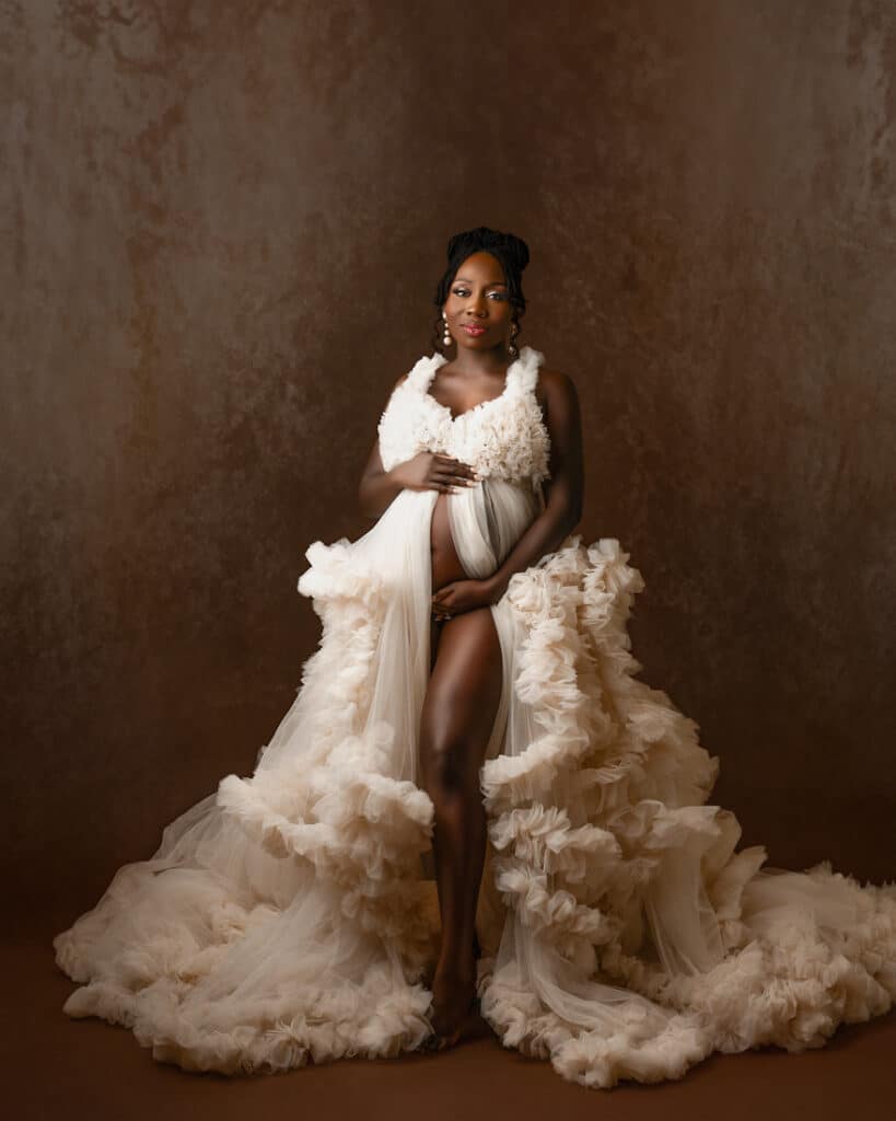 Woman Posed In Cream Gown Standing Against A Brown Textured Backdrop For Maternity Shoot