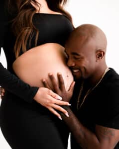 A Man Gently Kisses A Pregnant Woman'S Belly. Both Are Wearing Black Clothing Against A Plain White Background.