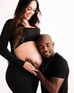 A Smiling Couple, Wearing Black Outfits, With The Man Resting His Head On The Woman'S Pregnant Belly.