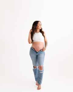 A Pregnant Woman In A White Top And Ripped Jeans Stands Barefoot Against A Plain Background, Looking Slightly To The Side.