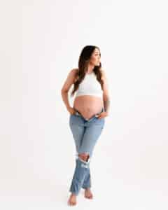Pregnant Person In A White Tank Top And Ripped Jeans Stands Barefoot Against A Plain White Background, Looking Off To The Side.