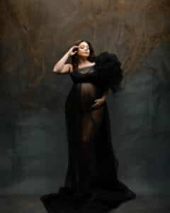 A Woman Stands In A Dark, Sheer Gown With A Voluminous Shoulder Detail, Hand Gently Resting On Her Forehead, Set Against A Textured, Earthy-Toned Background.