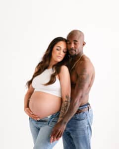 A Pregnant Woman In A White Tank Top And Jeans Stands Closely With A Shirtless Man In Jeans. Both Have Tattoos And Appear Calm.
