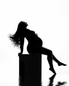 Silhouette Of A Pregnant Woman Sitting On A Box, Wearing A Form-Fitting Outfit. Her Long Hair Flows Down Her Back. The Scene Is Reflected In A Glossy Surface.