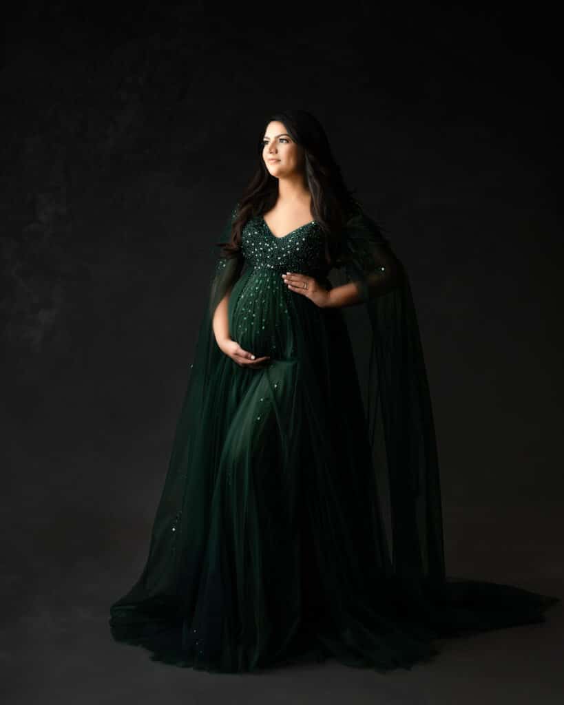 Woman In Green Flowing Gown Stands Against A Dark Background For Pregnancy Photos Dallas