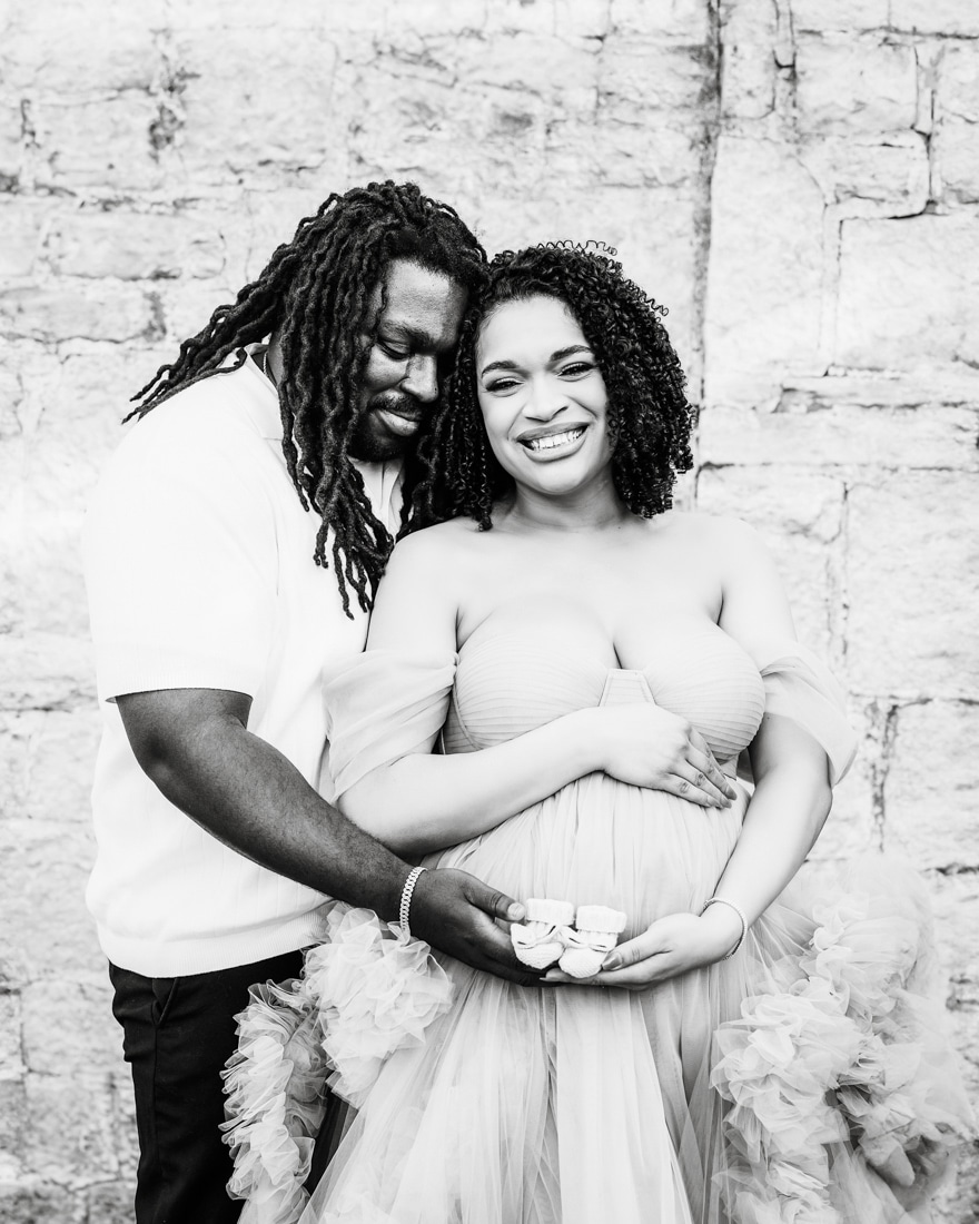 Couple Stands Against A Stone Wall With Man Sweetly Embracing For Dfw Maternity Photographer