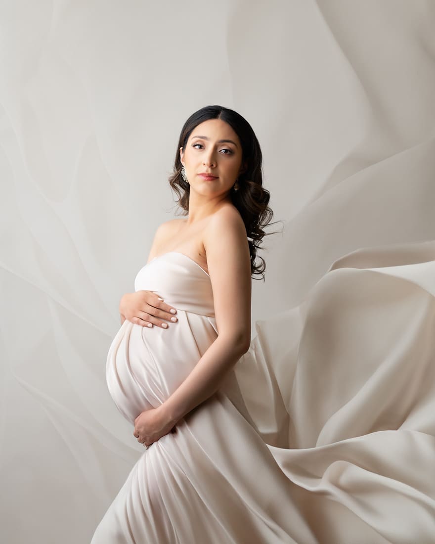 pregnant woman in flowing gown stands against a soft backdrop for dallas maternity photographer