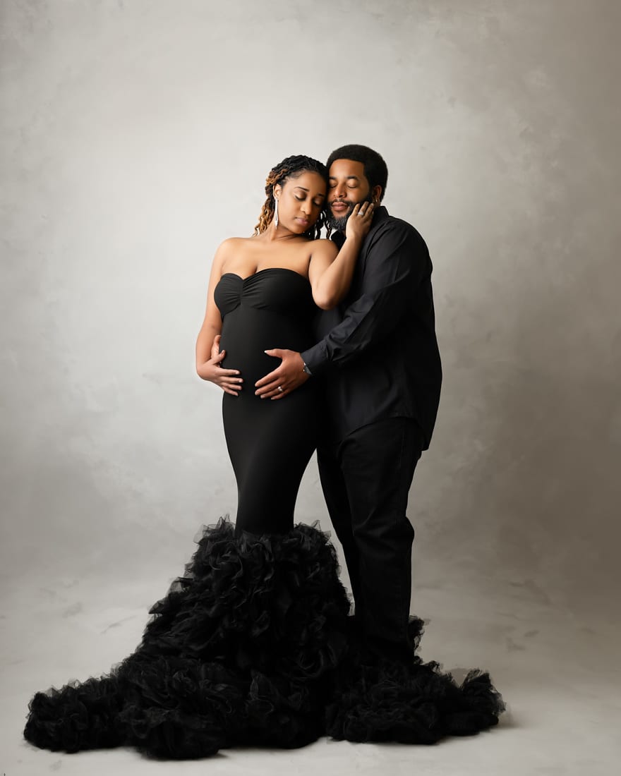 A Pregnant Woman In A Strapless Black Gown Stands Beside A Man In A Black Shirt; He Gently Touches Her Face As She Cradles Her Belly, Capturing A Tender Moment Of Love And Anticipation In Elegant Maternity Photography.