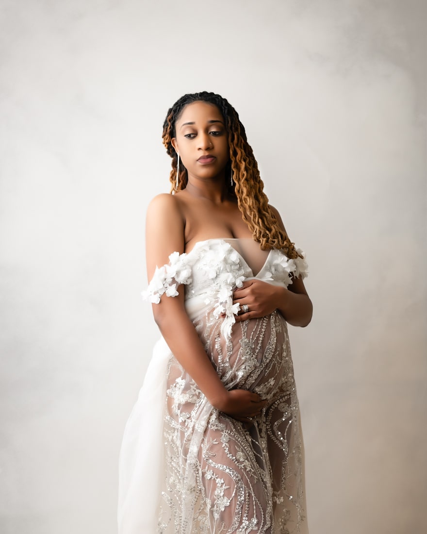 A Pregnant Woman In A Sheer, Embroidered Gown Stands Against A Light Background, Holding Her Baby Bump And Looking At The Camera—A Timeless Moment Of Maternity Photography.