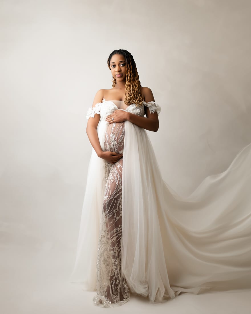 A Woman In A Sheer, Embellished Gown With A Flowing White Cape Stands In Front Of A Neutral Background, Holding Her Hands Over Her Midsection—A Graceful Moment Captured In Elegant Maternity Photography.