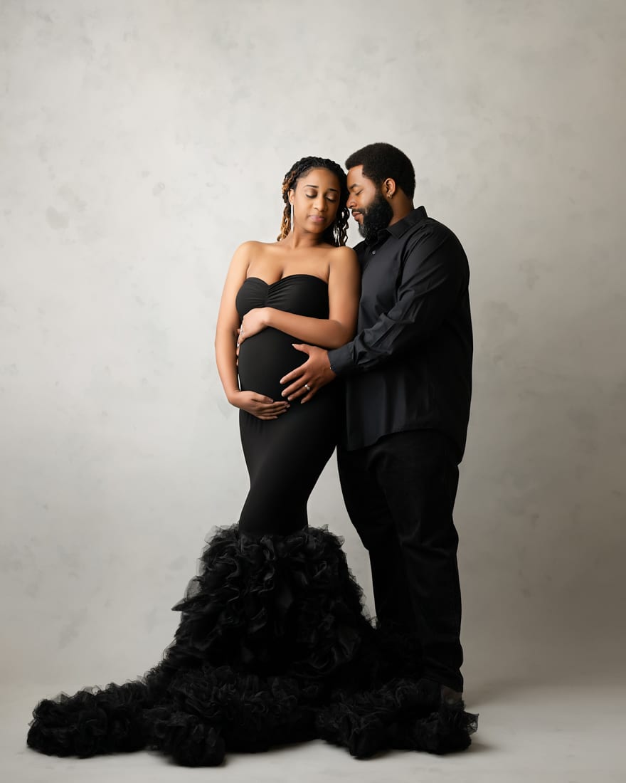 A Pregnant Woman In A Long Black Dress Stands Next To A Man In Black Clothing. Both Hold Her Belly And Gaze At Each Other, Capturing A Tender Moment Of Maternity Photography Against A Light, Plain Background.
