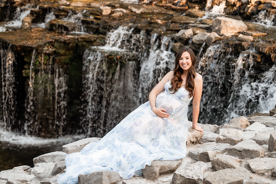 Woman In A Long, Light Blue Floral Dress Sits On Rocks In Smiling At The Camera For Dallas Maternity Photography