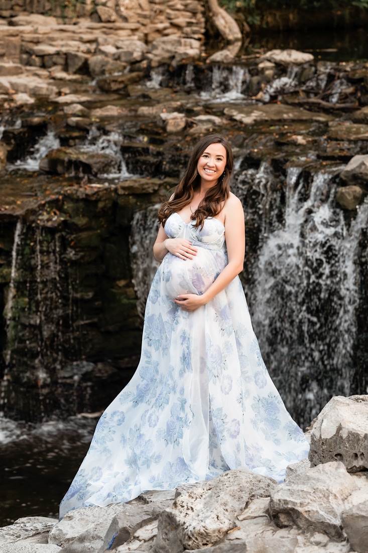 A Pregnant Woman In A Flowing, Light Blue Floral Dress Stands On Rocks In Front Of A Small Waterfall, Smiling At The Camera.