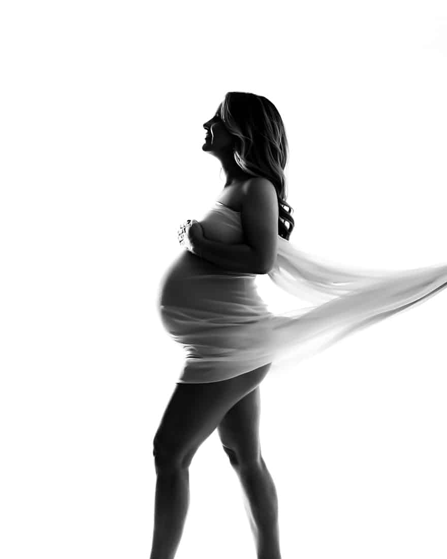 Silhouette Of A Pregnant Woman Standing In Profile, Draped In Flowing Fabric, Against A White Background.