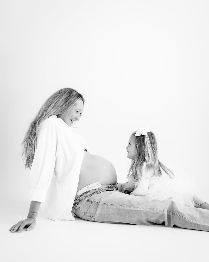 Pregnant Woman Sits On The Floor Smiling At A Young Daughter During A Maternity Photoshoot Dallas