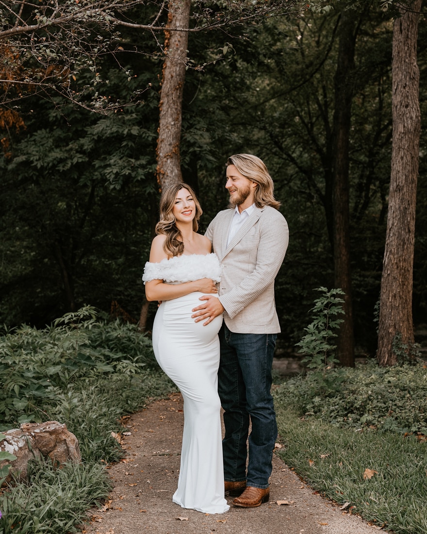 expecting couple smiling during outdoor maternity shoot in Dallas forest, woman in white off-shoulder gown holding baby bump