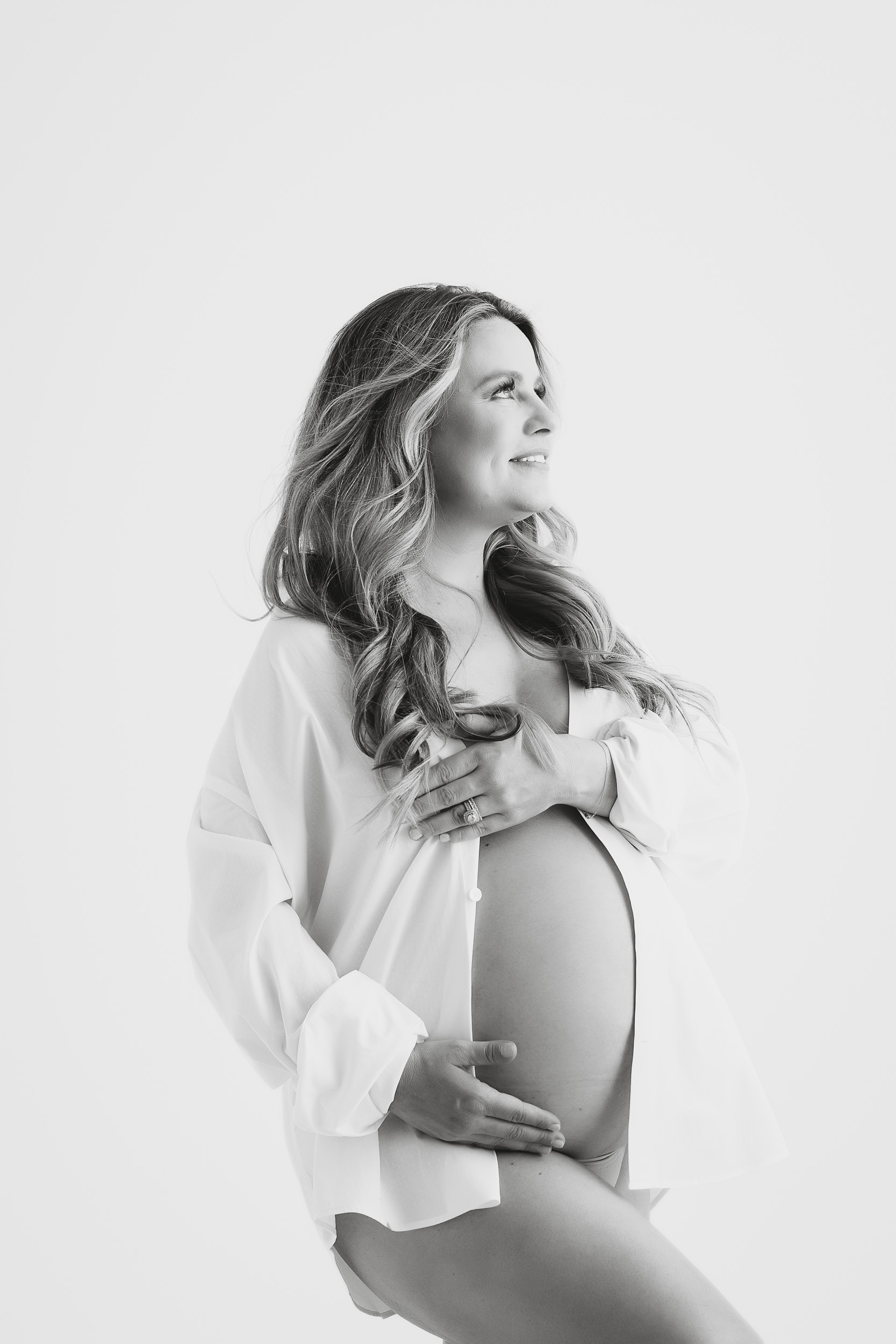 A Pregnant Woman In An Open White Shirt Poses In Profile During Her Maternity Session, Smiling And Looking Upward, With One Hand On Her Belly And The Other Covering Her Chest.