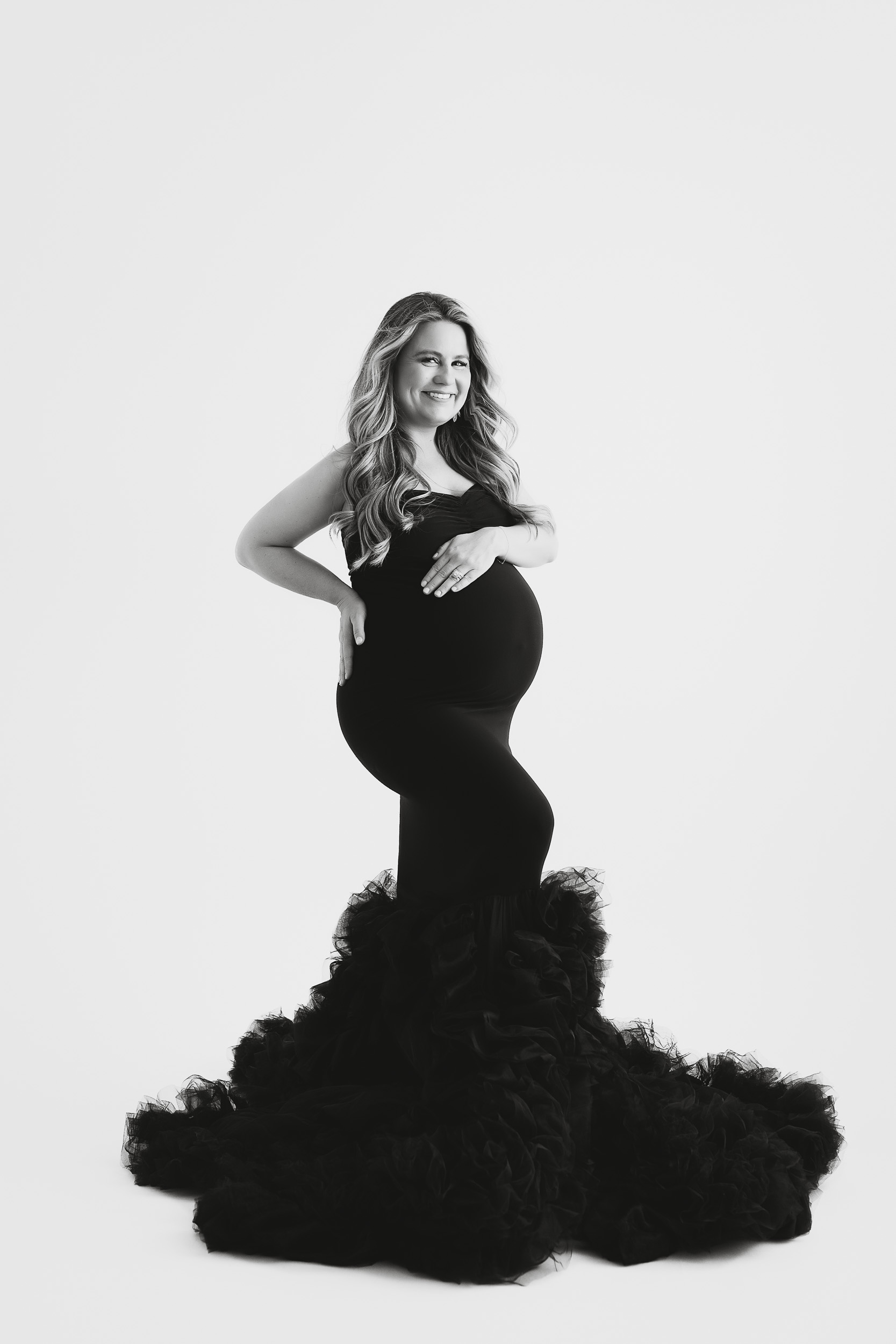A Pregnant Woman In A Fitted Black Gown With A Ruffled Train Poses And Smiles During Her Maternity Session In A Studio With A White Background.