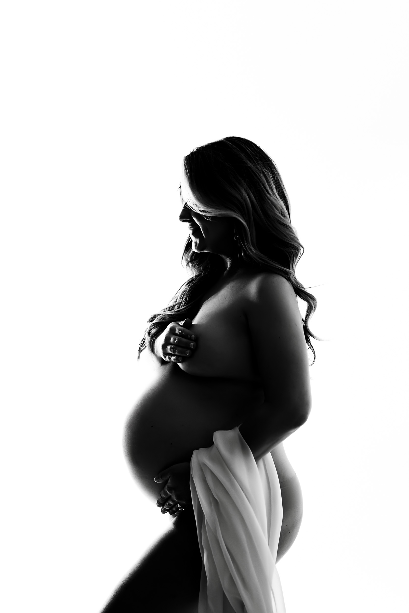 Black And White Side Profile Of A Pregnant Woman During A Maternity Session, Partially Nude, Covering Her Chest With One Hand And Draped In Light Fabric, Set Against A White Background.
